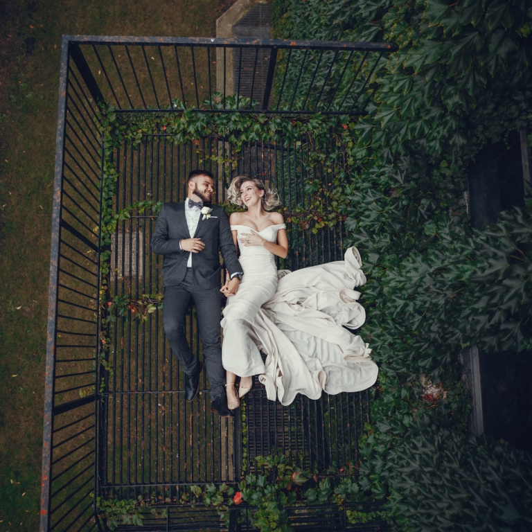 Beautiful bride in a long white dress. Handsome groom in a black suit. Couple lie on a metal