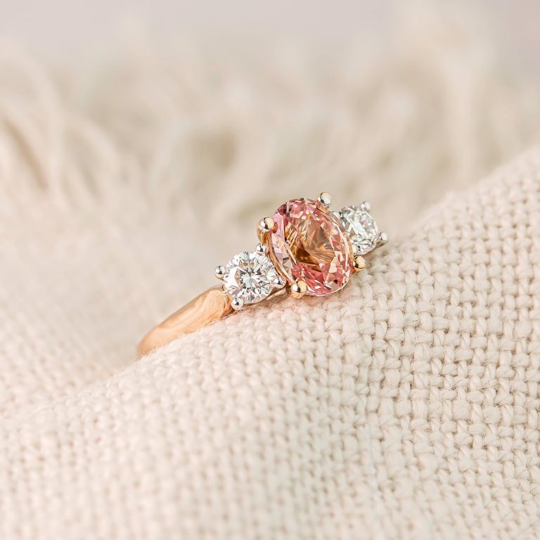 Three Stone Ring Featuring an Oval Padparadscha Sapphire and Two Round Brilliant Cut Diamonds