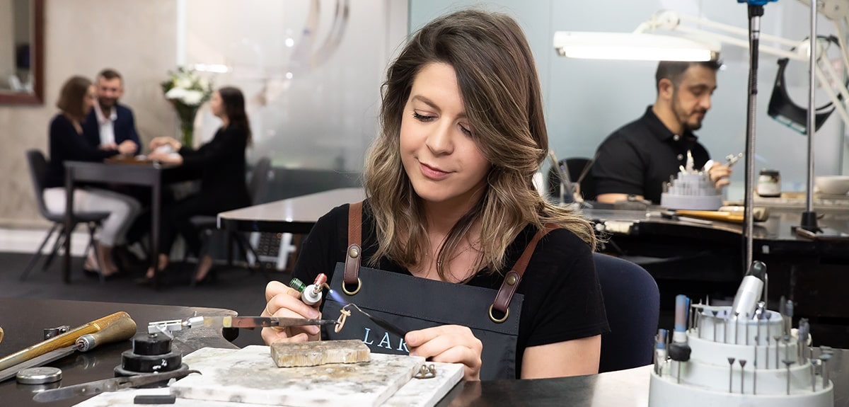 A jeweller in the Larsen Studio crafting a custom engagement ring. Customers meet with a design consultant in the background